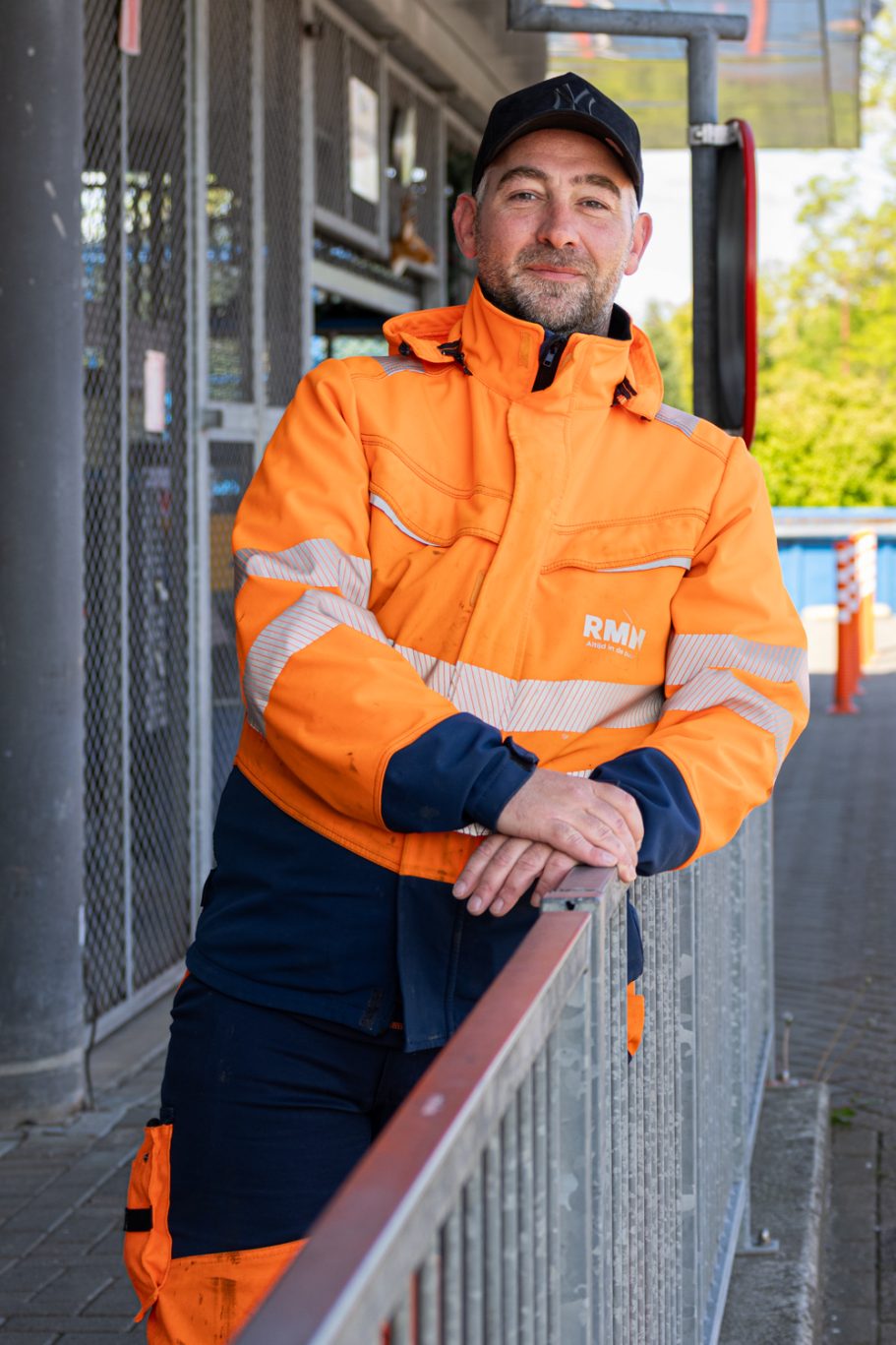 Man in oranje beschermkleding leunt op een railing, met een cap op in een stedelijke omgeving.